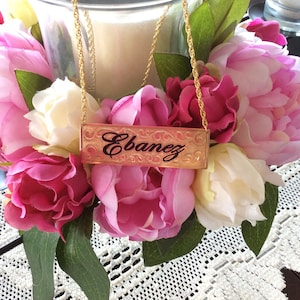 May include: A gold chain necklace with a rectangular nameplate pendant that reads "Ebanez". The necklace is displayed on a table with pink and white flowers.