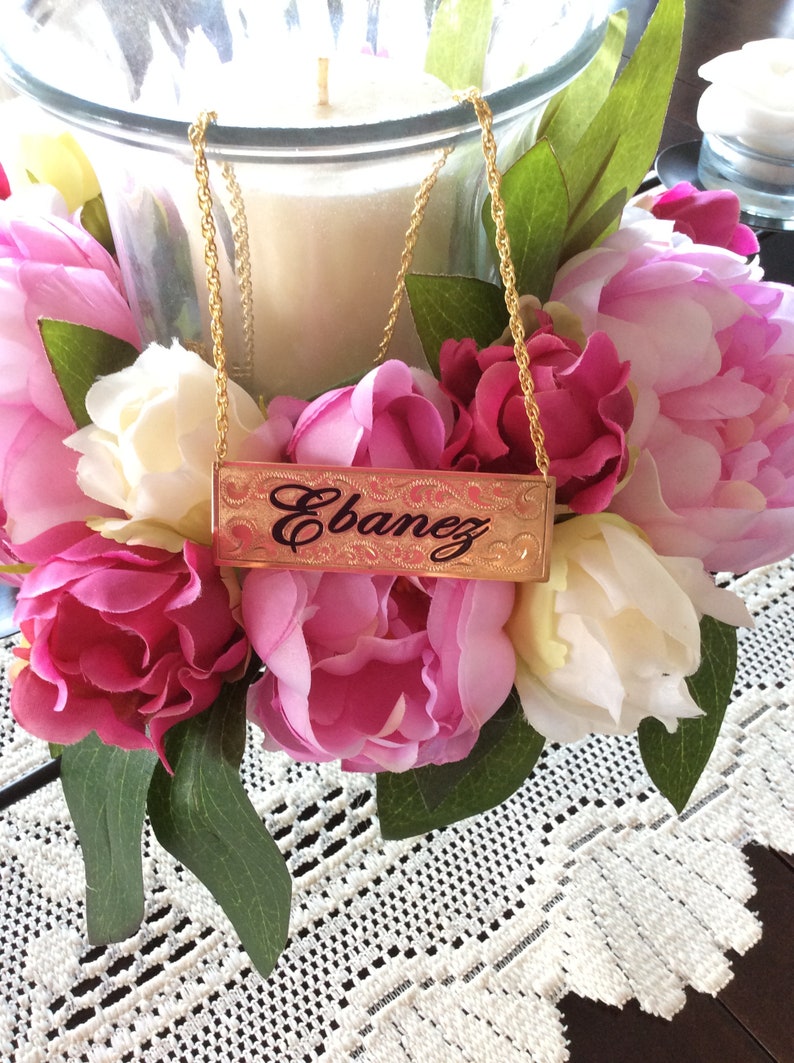 May include: A gold chain necklace with a rectangular nameplate pendant that reads "Ebanez". The necklace is displayed on a table with pink and white flowers.