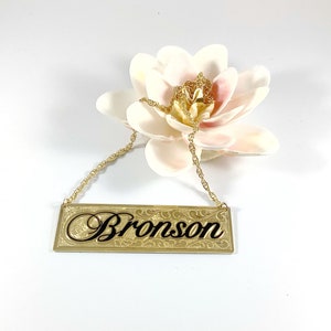 May include: Gold necklace with a rectangular nameplate pendant that says "Bronson" in black script. The pendant has a decorative border.
