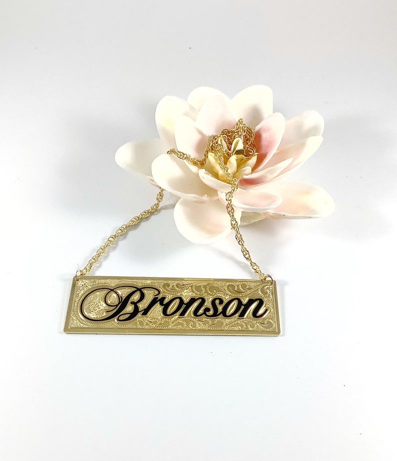 May include: Gold necklace with a rectangular nameplate pendant that says "Bronson" in black script. The pendant has a decorative border.
