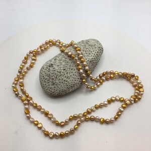 K&ouml;nnte beinhalten: Eine lange Halskette aus zwei Reihen Perlen. Die Perlen sind in einem Mix aus hellrosa und goldfarben. Die Kette liegt auf einem Stein.