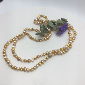 K&ouml;nnte beinhalten: Eine lange Halskette aus goldenen und cremefarbenen Perlen. Die Kette liegt auf einer wei&szlig;en Oberfl&auml;che mit einem Zweig getrockneten Eukalyptus und lila Bl&uuml;ten.