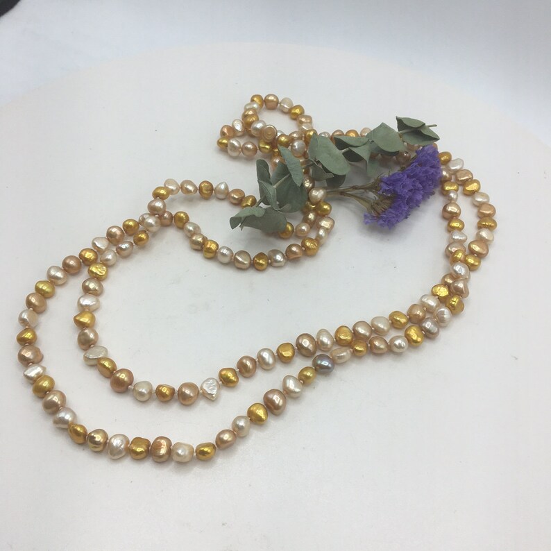 K&ouml;nnte beinhalten: Eine lange Halskette aus goldenen und cremefarbenen Perlen. Die Kette liegt auf einer wei&szlig;en Oberfl&auml;che mit einem Zweig getrockneten Eukalyptus und lila Bl&uuml;ten.