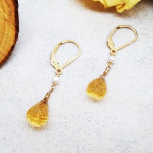 May include: A pair of gold earrings with a teardrop-shaped yellow gemstone and a small white pearl on each earring.