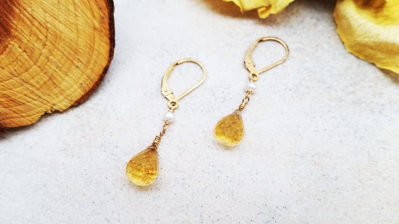 May include: A pair of gold earrings with a teardrop-shaped yellow gemstone and a small white pearl on each earring.