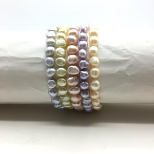 May include: A set of six pearl bracelets in various pastel colours, including lavender, pink, and cream. The bracelets are made of small, round pearls and are stacked on top of each other.