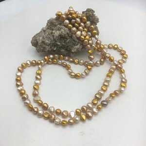 K&ouml;nnte beinhalten: Eine lange Perlenkette mit einem Mix aus goldenen und cremefarbenen Perlen.