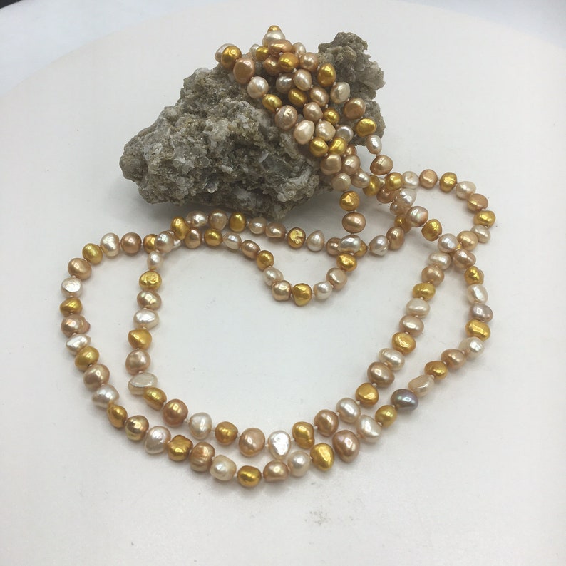 K&ouml;nnte beinhalten: Eine lange Perlenkette mit einem Mix aus goldenen und cremefarbenen Perlen.