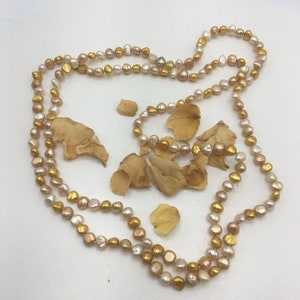 K&ouml;nnte beinhalten: Eine lange Halskette aus goldenen und cremefarbenen Perlen. Die Kette liegt auf einer wei&szlig;en Oberfl&auml;che mit getrockneten Bl&uuml;tenbl&auml;ttern.