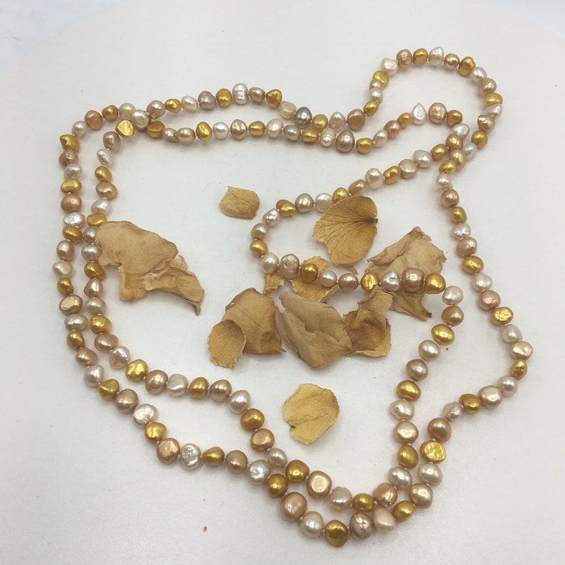 K&ouml;nnte beinhalten: Eine lange Halskette aus goldenen und cremefarbenen Perlen. Die Kette liegt auf einer wei&szlig;en Oberfl&auml;che mit getrockneten Bl&uuml;tenbl&auml;ttern.
