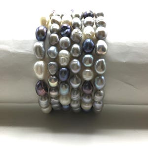 May include: A multi-strand bracelet featuring irregularly shaped pearls in shades of white, grey, and dark purple. The pearls are strung together, creating a textured and elegant accessory.