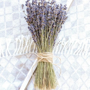Könnte beinhalten: Ein Bund getrockneter Lavendel mit lila Blüten und grünen Stielen, zusammengebunden mit Bindfaden. Das Bouquet liegt auf einem weiß-grau gemusterten Stoff und erzeugt eine rustikale und natürliche Ästhetik. Lavendel wird oft für Aromatherapie und Dekoration verwendet.