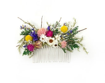 May include: A white hair comb decorated with dried flowers. The flowers are pink, yellow, white, and purple. The comb is a great accessory for weddings or special occasions.