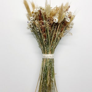 Könnte beinhalten: Ein Trockenblumenstrauß mit weißen und braunen Blumen und Gräsern, gebunden mit einem weißen Band.