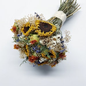 Könnte beinhalten: Ein Trockenblumenstrauß mit Sonnenblumen, weißen und gelben Wildblumen und blauem Lavendel. Der Strauß ist mit einem weißen Spitzenband gebunden.