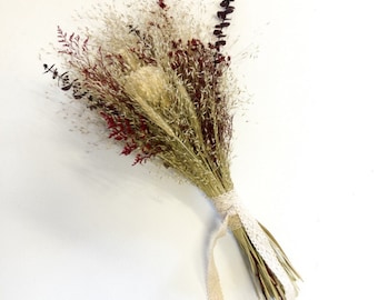 May include: A dried flower bouquet with a mix of beige, brown, and red dried grasses and flowers tied together with a white lace ribbon.