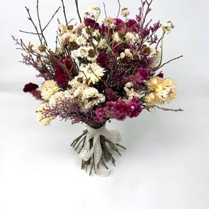 Puede incluir: Un ramo de flores secas con siemprevivas de color crema, celosía de color magenta intenso y ramitas marrones finas. Los tallos están agrupados y atados con una cinta de encaje blanco, creando un arreglo texturizado de elementos botánicos secos.