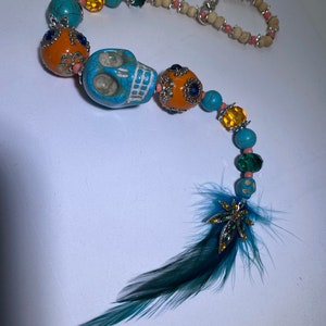 May include: A turquoise and orange beaded necklace with a turquoise skull charm and a green feather with a silver charm at the end.