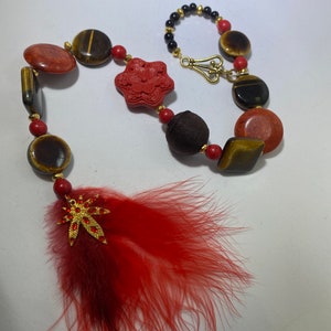May include: A red and brown beaded necklace with a gold clasp and a red feather charm. The beads are made of tiger's eye, coral, and wood.
