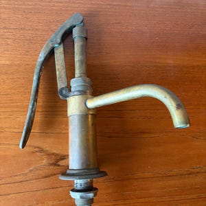 May include: Vintage brass faucet with a lever handle and curved spout. The faucet has a cylindrical body and a threaded base. The brass has a patina, showing signs of age and use. The faucet is designed for water dispensing.