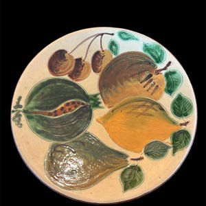 May include: A round ceramic plate with a hand-painted design of fruit. The plate features a pear, a lemon, a pomegranate, and cherries. The fruit is painted in shades of green, yellow, brown, and red.