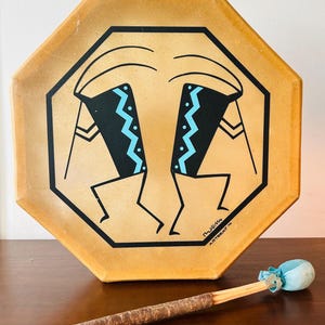 Puede incluir: Un tambor octogonal de color beige con un diseño negro y azul de dos figuras. El tambor viene con un palo de madera con una cabeza de fieltro azul.