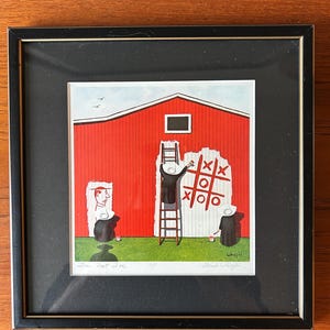 Puede incluir: Impresión enmarcada que representa un granero rojo con un juego de tres en raya en un lateral. Se muestran tres figuras, una en una escalera, otra sosteniendo un papel y una tercera cerca del juego. La impresión está firmada e incluye el texto "See Dot Doe".