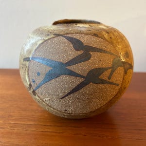 May include: A round, speckled ceramic vase with a small opening at the top. The vase features a textured, tan background with a circular design of stylized, dark blue birds in flight. The vase is approximately 5 inches in diameter.