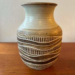 Könnte beinhalten: Eine Keramikvase mit hellbeigem Oberteil und braunem Boden. Die Vase hat horizontale, wellenförmige Bänder mit einem weißen, linearen Muster. Die Vase hat einen abgerundeten Körper und einen ausgestellten Rand, geeignet für die Inneneinrichtung.