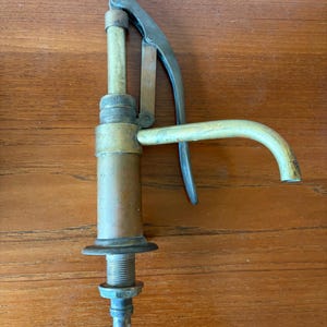 May include: An antique brass and copper water pump with a curved spout and a lever handle. The pump has a threaded base and a cylindrical body. The metal has a weathered appearance, suggesting age and use. The pump is likely for a well or other water source.