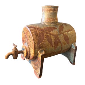 Puede incluir: Un jarrón de cerámica con forma de barril con un pico y patas. El jarrón está decorado con un esmalte marrón y amarillo y tiene un diseño de hojas.