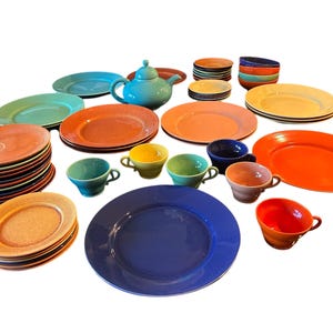 Peut inclure: Un ensemble de vaisselle vintage en céramique colorée, comprenant des assiettes, des bols, des tasses et une théière. L'ensemble présente une variété de couleurs vives, notamment l'orange, le jaune, le bleu, le vert et le turquoise.
