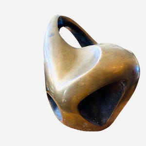 May include: A bronze-colored, abstract sculpture with a rounded form and a handle-like opening at the top. The sculpture has several geometric cutouts and a textured surface, suggesting an aged or weathered appearance. The overall design is modern and artistic.