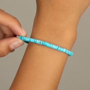 May include: A turquoise beaded bracelet is shown on a wrist. The bracelet is made of small, round, light blue beads strung together. The bracelet is being put on the wrist. The background is a solid, neutral color.
