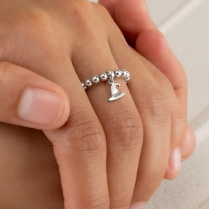 May include: A silver beaded ring with a small charm shaped like a coffee cup. The ring is on a hand, with a light-colored background. The ring is made of small, round beads and the charm hangs from the center.