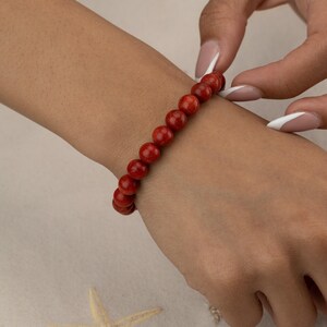 May include: A red beaded bracelet with round beads is worn on a wrist. The bracelet is made of small, evenly sized beads. The skin tone is light. A hand is adjusting the bracelet. The background is a light beige color.