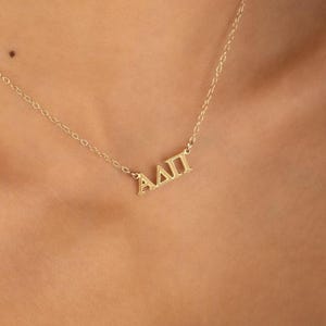 May include: A gold-toned necklace featuring a delicate chain and a pendant displaying the Greek letters Alpha, Delta, and Pi in a polished finish. The pendant is centered on the chain, which connects to the letters at both ends, creating a simple design.