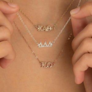 May include: Three layered necklaces featuring Greek letter pendants. The top necklace is gold-toned with the letters KAO, the middle is silver-toned with three delta symbols, and the bottom is gold-toned with the letters XO. Each has a delicate chain.