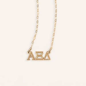 May include: A gold-toned necklace featuring a delicate chain and a pendant displaying three Greek letters: Alpha, Theta, and Delta. The letters are connected in a horizontal row, creating a polished metallic charm suspended from the fine link chain.