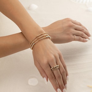 May include: A set of three gold-toned bracelets featuring smooth round beads and a twisted texture, worn alongside a stack of three gold-toned rings with small round beads. The jewellery pieces are polished and have a reflective metallic finish.