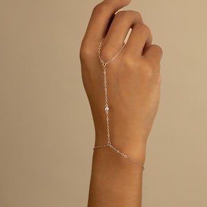 May include: A delicate silver-toned hand chain featuring a thin metal link chain that connects a ring loop to a wrist bracelet. The design includes a single small, clear, round-cut stone set in the center of the chain segment between the wrist and finger.