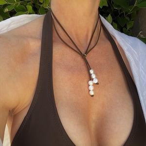 Puede incluir: Un collar de cordón de cuero marrón con un racimo de perlas blancas que cuelgan de él. El collar lo lleva una persona con un top de bañador marrón.