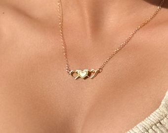 May include: A gold-toned necklace featuring a central pendant composed of three interconnected heart shapes. The center heart is solid, while the two flanking hearts are open-work outlines. The pendant is suspended from a delicate gold-toned chain.