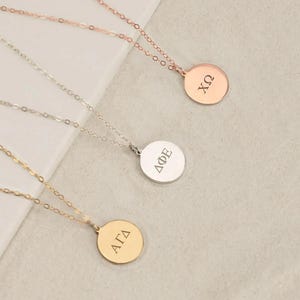 Könnte beinhalten: Drei Halsketten mit dünnen Metallketten und runden Anhängern. Der oberste Anhänger ist aus Roségold mit den griechischen Buchstaben XΩ, der mittlere aus Silber mit ΔΦΕ und der untere aus Gold mit ΑΓΔ. Jeder Anhänger hat eine glatte Oberfläche.