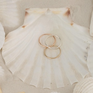 May include: Three thin gold rings stacked on top of each other on a white seashell. The rings are different sizes and are made of gold, silver, and rose gold.