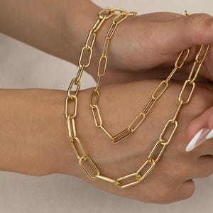 May include: A gold-colored chain necklace and bracelet set. The necklace features a series of elongated oval links. The bracelet is a matching design. The jewelry is displayed on a person's hand.
