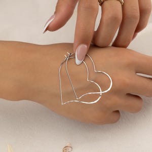 May include: Silver heart-shaped hoop earrings. The earrings are held against a light skin tone, with a gold ring stack on the finger. The background is a light, neutral color.