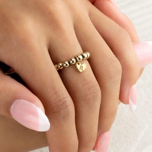 Puede incluir: Un anillo dorado con cuentas y un dije en forma de corazón. El dije tiene la letra "R". El anillo está en un dedo.