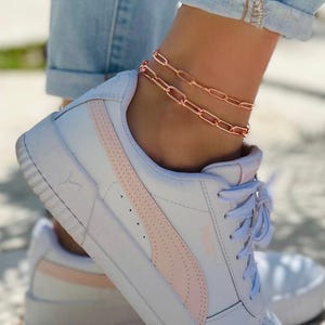 May include: A rose gold ankle chain with two rows of linked oval shapes. The chain is worn on a person's ankle above white sneakers with a pink stripe.
