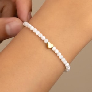 Puede incluir: Pulsera de cuentas blancas con un dije de corazón dorado. La pulsera está hecha de pequeñas cuentas blancas redondas y presenta un solo dije de corazón dorado en el centro. La pulsera se muestra en una muñeca.